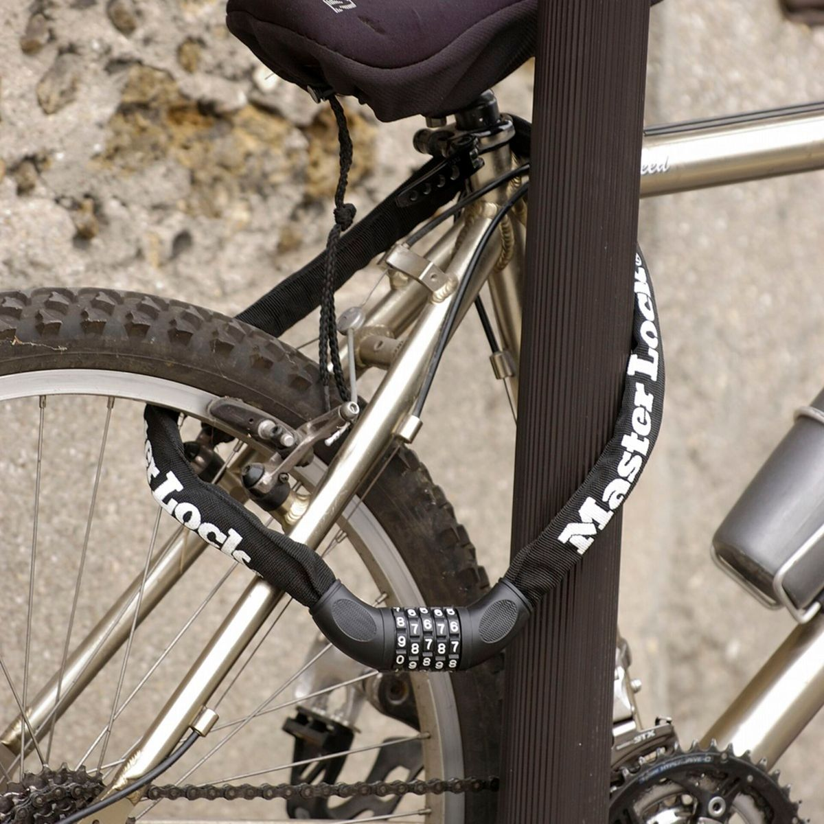 Ein silbernes Fahrrad, das mit einem schwarzen Stangen mit einem schwarzen Master Lock-Kombinationsschloss gesichert ist. Das Fahrrad steht in der Nähe einer verwitterten Steinmauer.