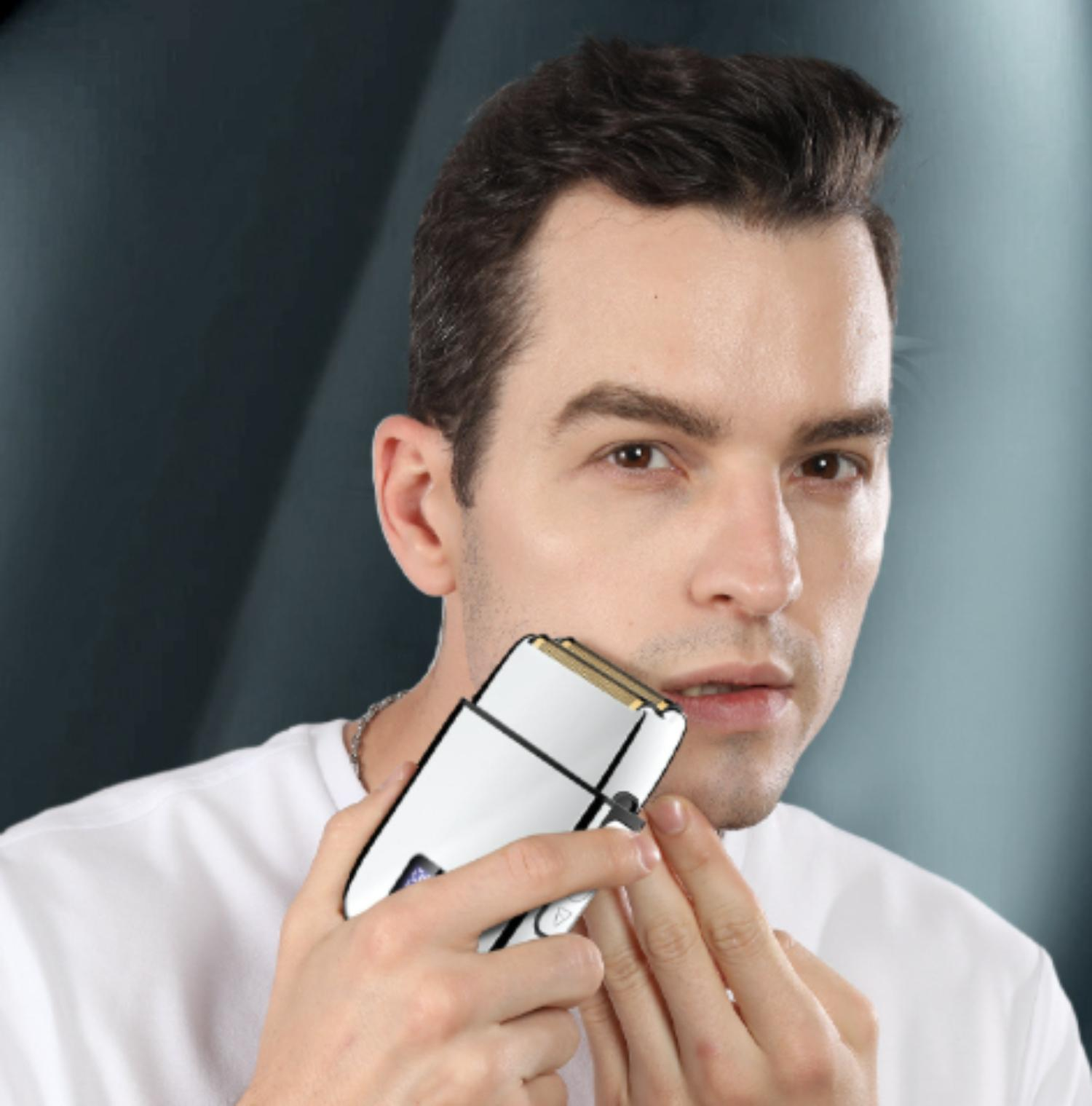 Ein Mann rasiert sich mit einem Elektrorasierer. Er trägt ein weißes Hemd vor einem dunklen Hintergrund. Er schaut in die Kamera.