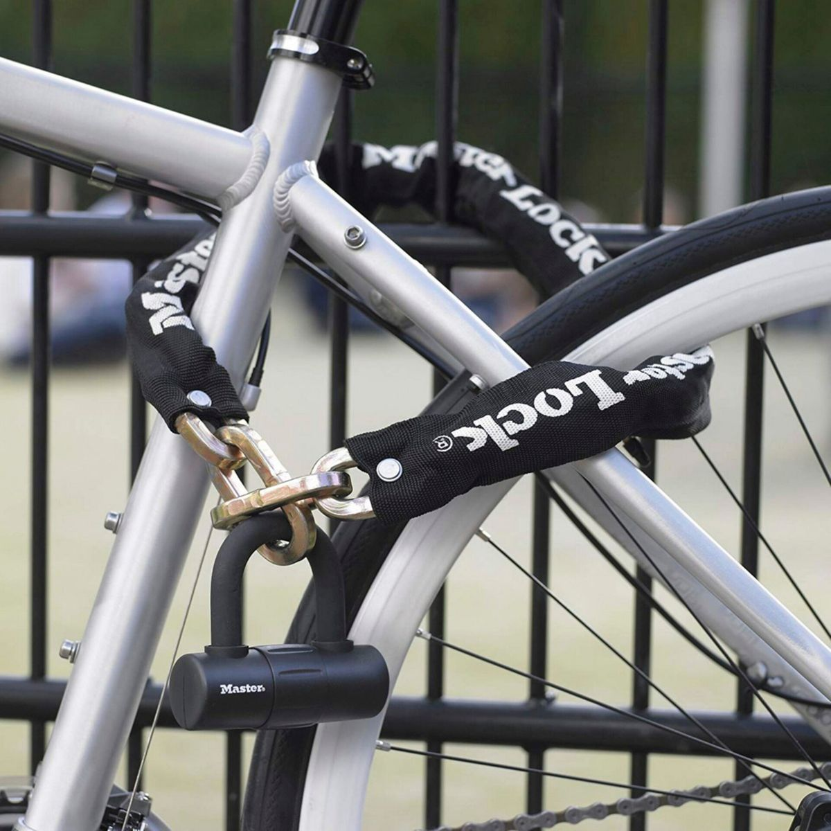 Ein silbernes Fahrrad ist mit einer schwarzen Master Lock Kette und einem Vorhängeschloss an einem schwarzen Zaun befestigt.