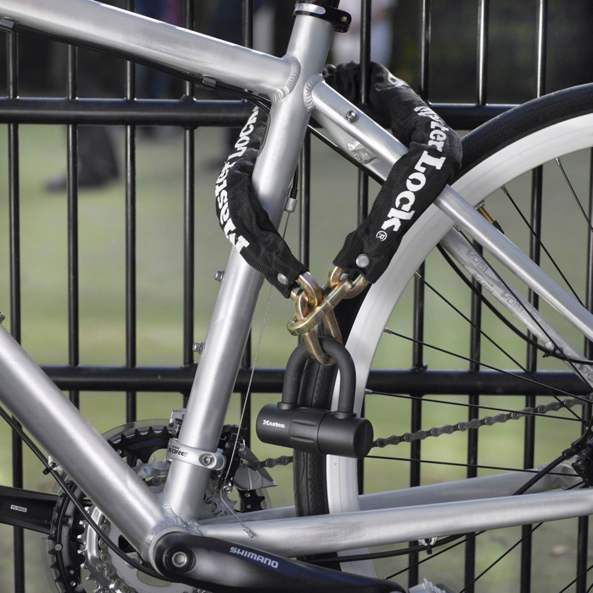 Ein silbernes Fahrrad ist mit einer schwarzen Kette und einem Master Lock an einem schwarzen Zaun befestigt. Der Hinterreifen ist weiß. Grünes Gras ist im Hintergrund.