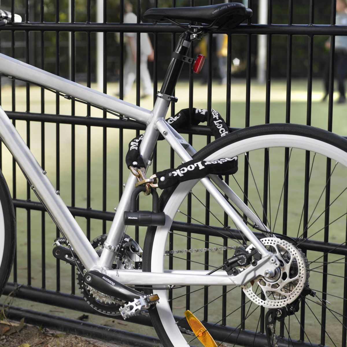 Silbernes Fahrrad, das an einem schwarzen Zaun angeschlossen ist. Ein schwarzes Kettenschloss sichert das Fahrrad. Menschen gehen im Hintergrund.