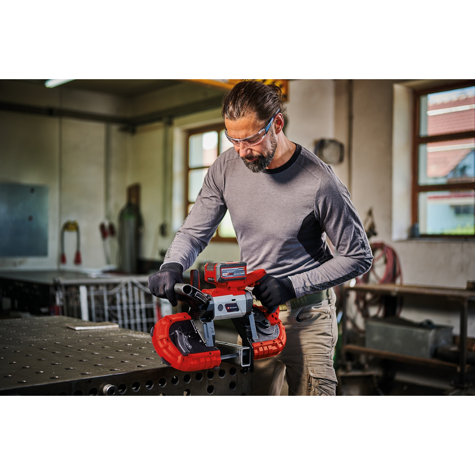 Ein Mann benutzt eine rot-silberne Bandsäge in einer Werkstatt. Er trägt eine Schutzbrille und Handschuhe und arbeitet an einem Metalltisch.
