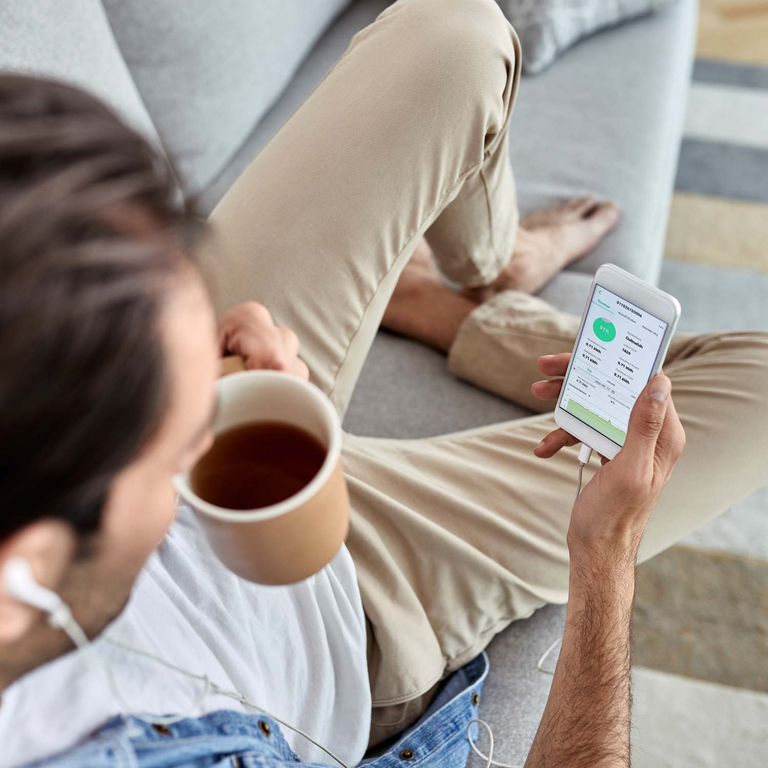Una persona sentada en un sofá, sosteniendo una taza de té y un teléfono con datos. Viste pantalones beige y una camisa blanca, con auriculares.