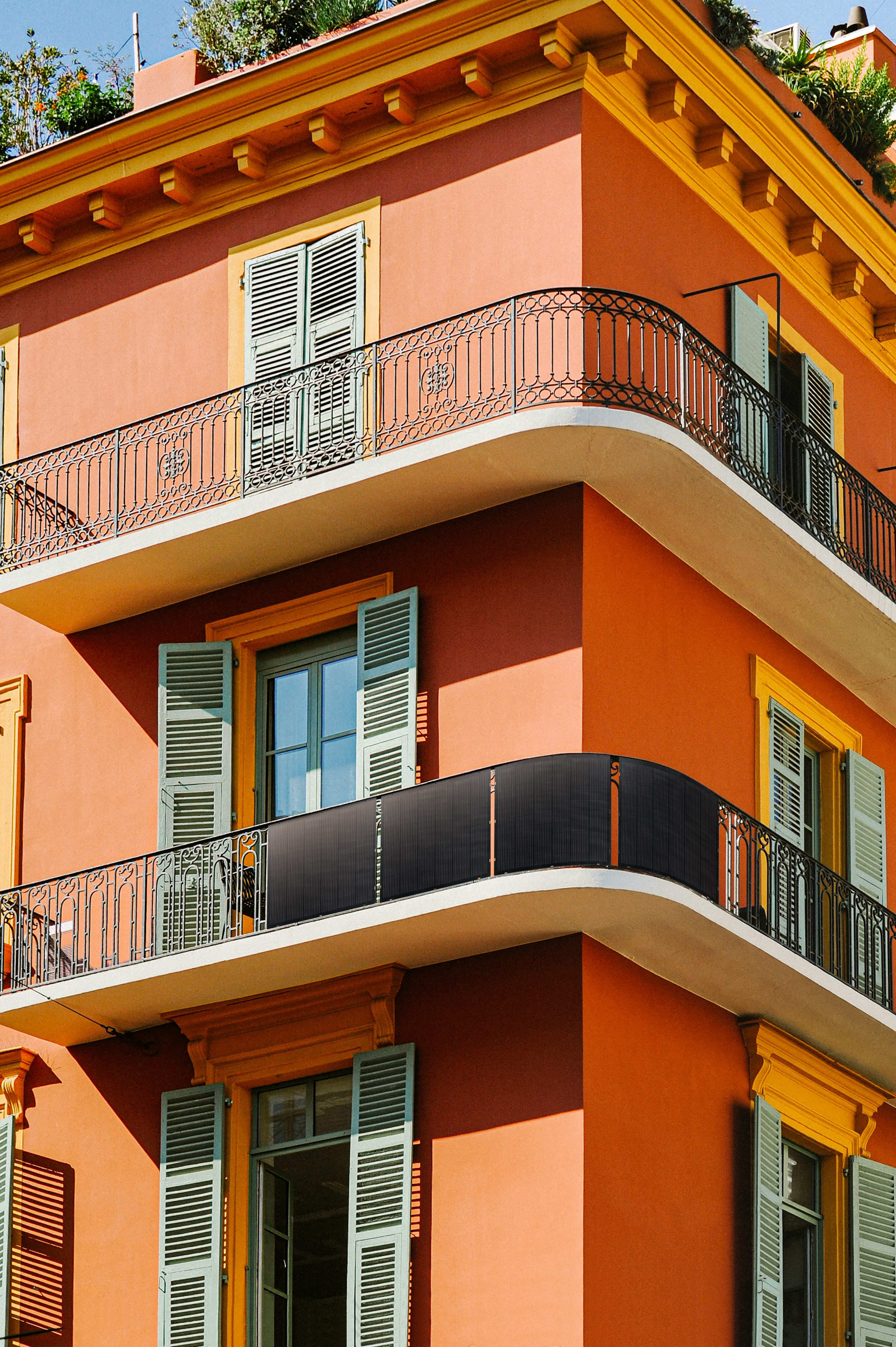 Esquina de un edificio con balcones, contraventanas y una valla negra. El edificio es naranja con molduras amarillas y contraventanas azules.