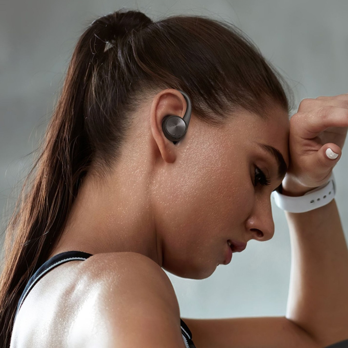 Frau mit Kopfhörern im Ohr, Hand an der Stirn. Sie trägt einen Fitnesstracker und ein schwarzes Oberteil. Schweiß sichtbar.