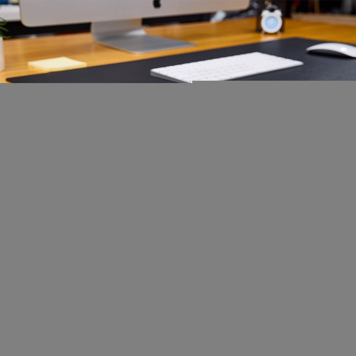 Biurko z komputerem, klawiaturą, myszą i budzikiem. Żółta karteczka samoprzylepna leży na biurku.