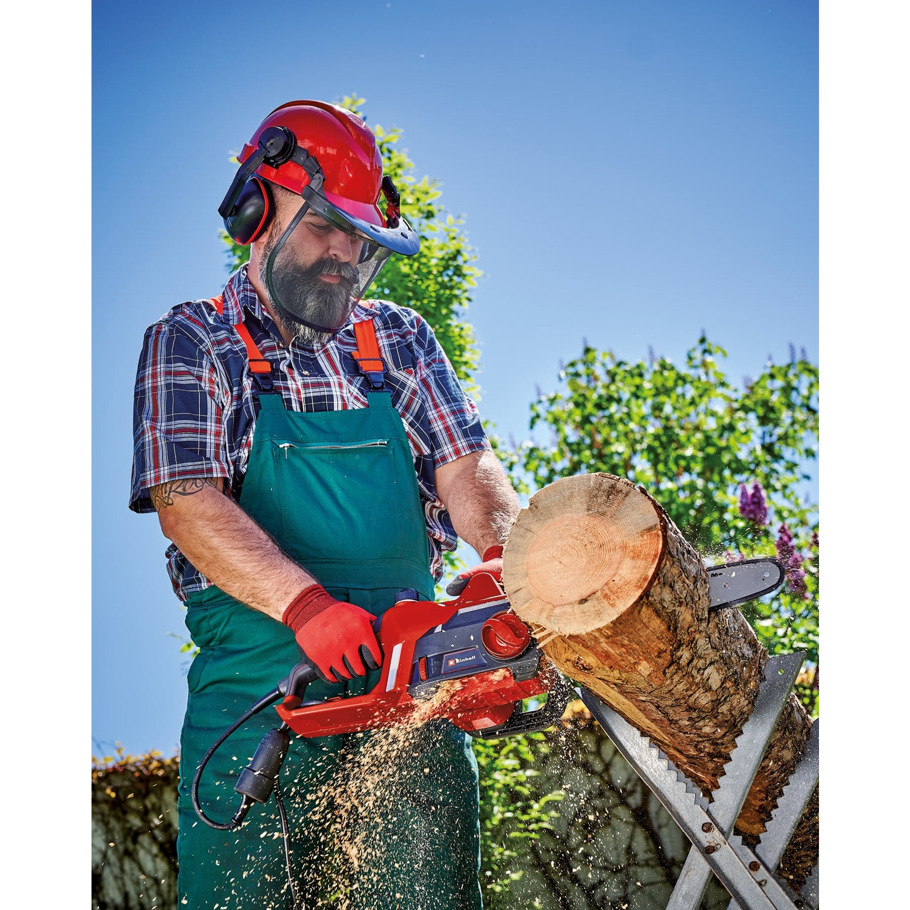 Mężczyzna w sprzęcie ochronnym tnie kłodę piłą łańcuchową. Wióry lecą. Błękitne niebo i zielone liście w tle.
