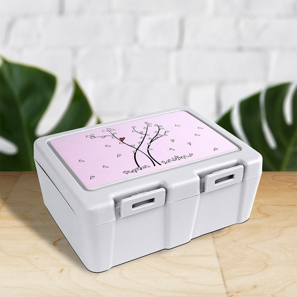 Una lonchera blanca con tapa rosa. La tapa tiene un diseño de árbol con corazones. Está sobre una mesa de madera con fondo borroso.