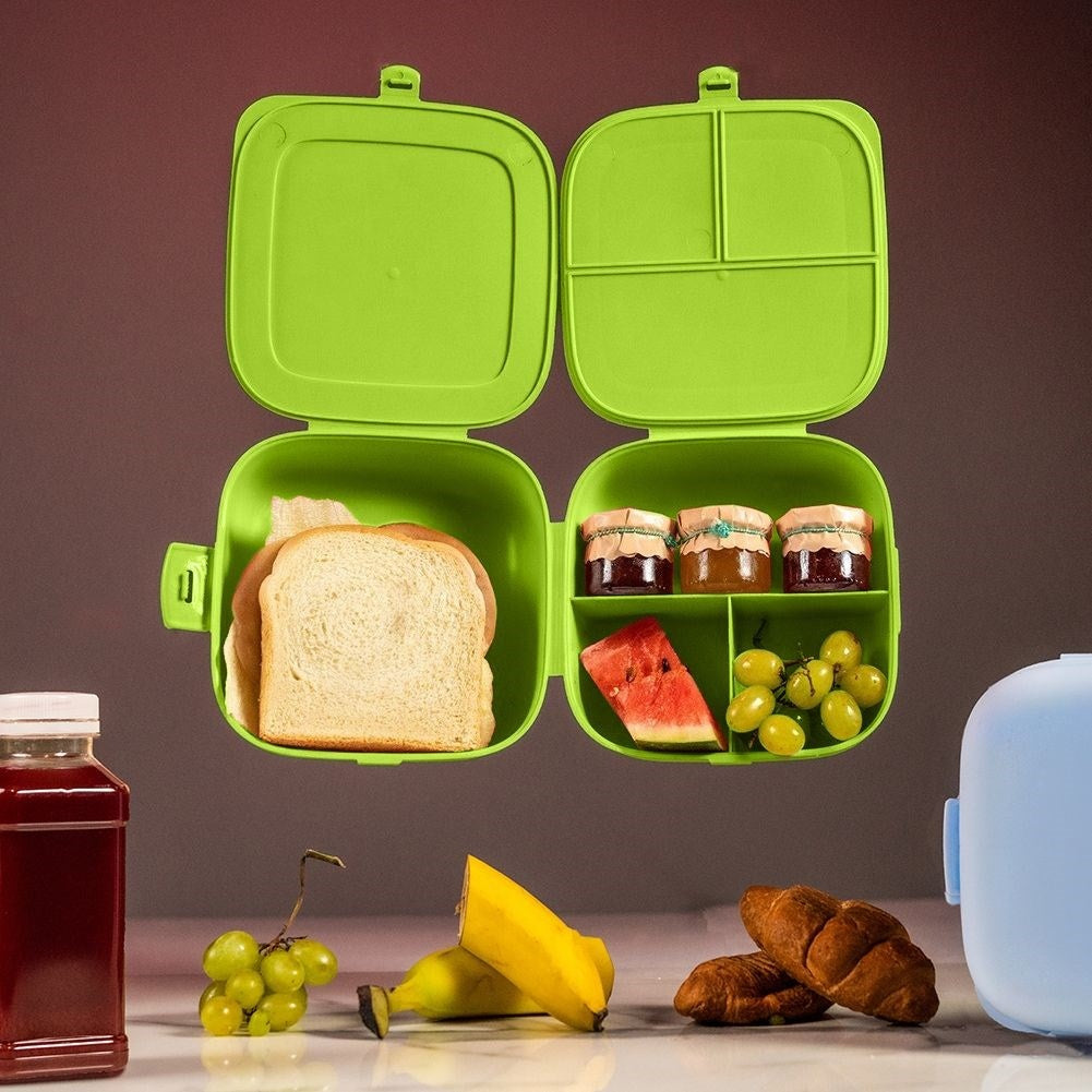 Una lonchera verde abierta, con pan, fruta y mermelada. Uvas, plátanos, cruasanes y una botella de zumo también en la mesa.