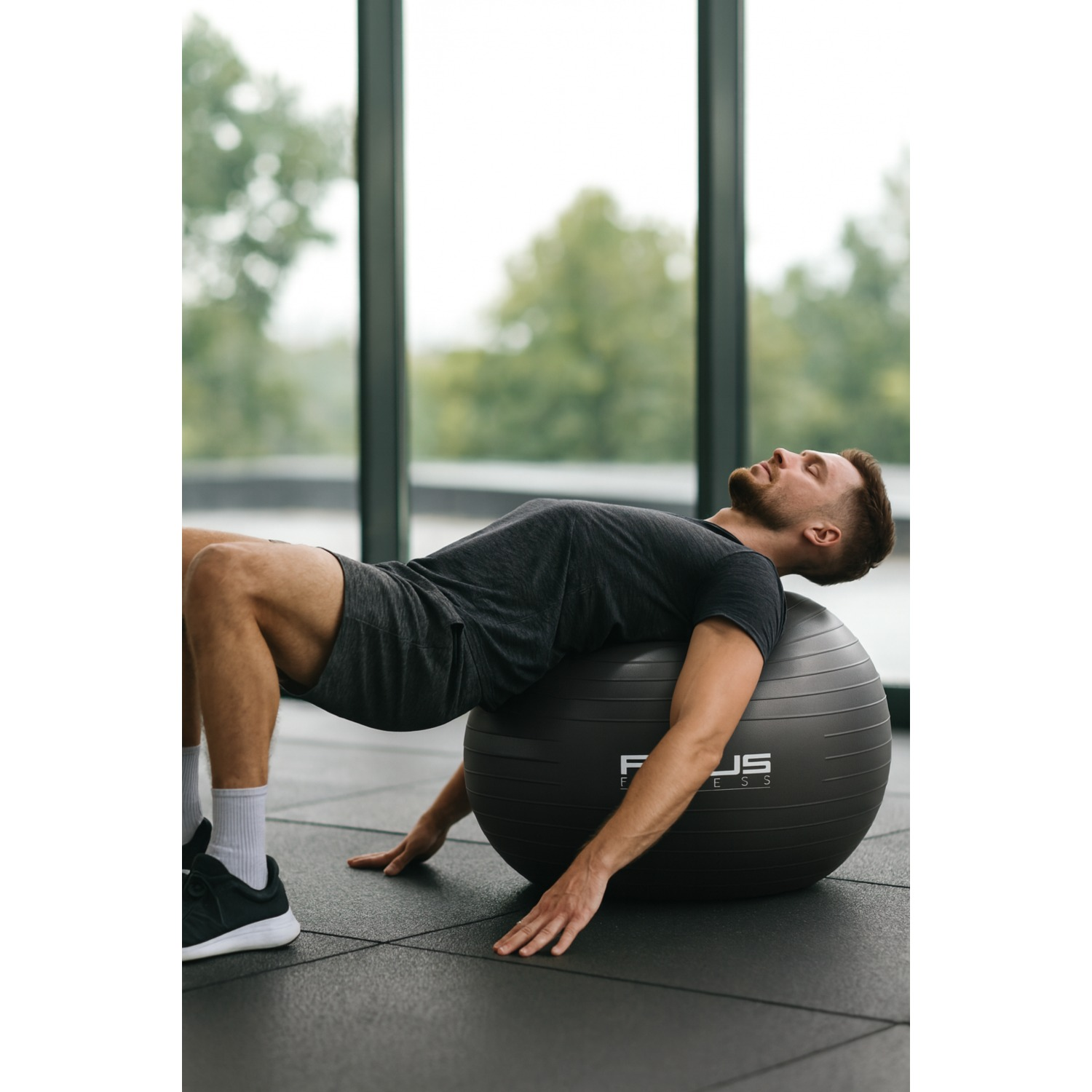 Un homme est allongé sur un ballon de fitness dans une salle de sport. Il porte un t-shirt sombre et un short. Le ballon est noir, et il repose sa tête dessus.