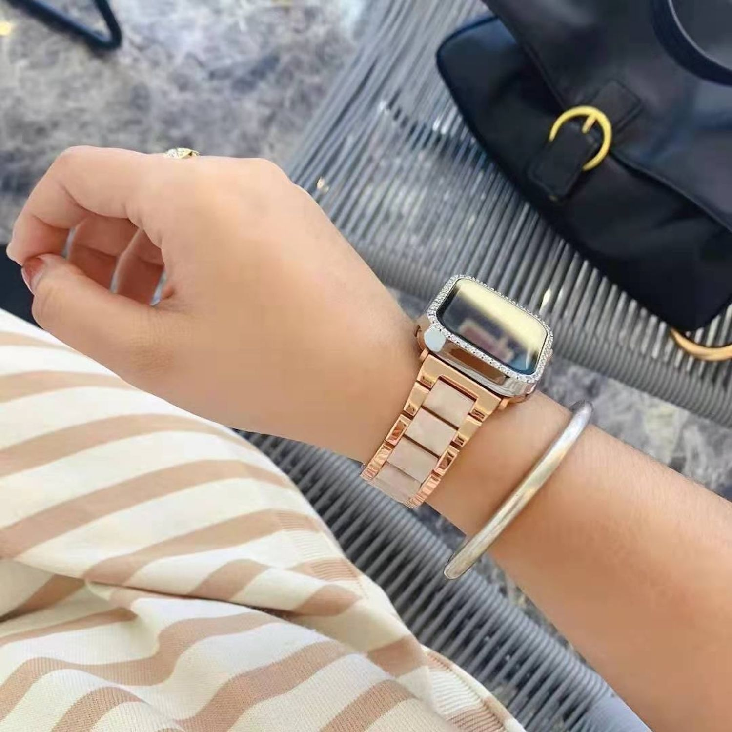 Eine Person, die eine roségoldene Uhr mit weißem Armband und silbernem Armband trägt. Im Hintergrund ist eine schwarze Handtasche zu sehen.
