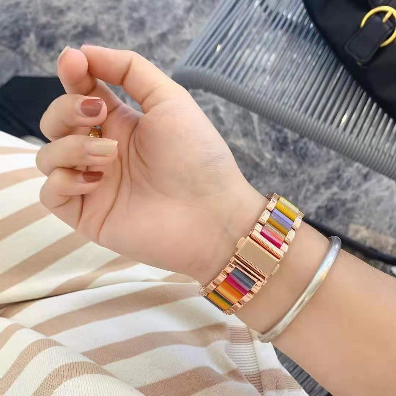 Eine Hand mit lackierten Nägeln trägt eine bunte Uhr und ein silbernes Armband. Im Hintergrund eine schwarze Handtasche mit goldenem Verschluss.