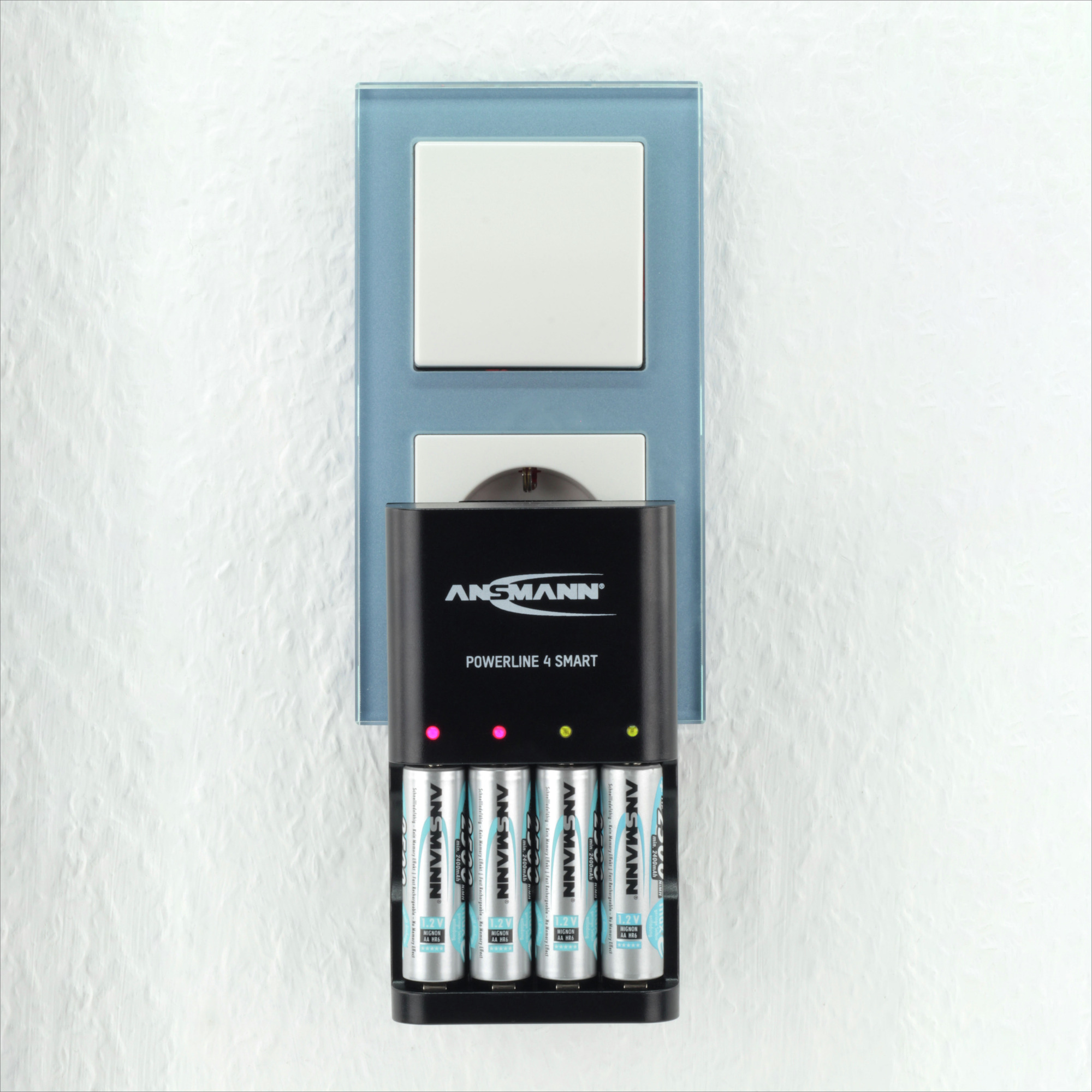 Eine Wandsteckdose mit Ladegerät und vier AA-Batterien. Das Ladegerät ist schwarz mit der Marke ANSMANN. Die Wand ist weiß.