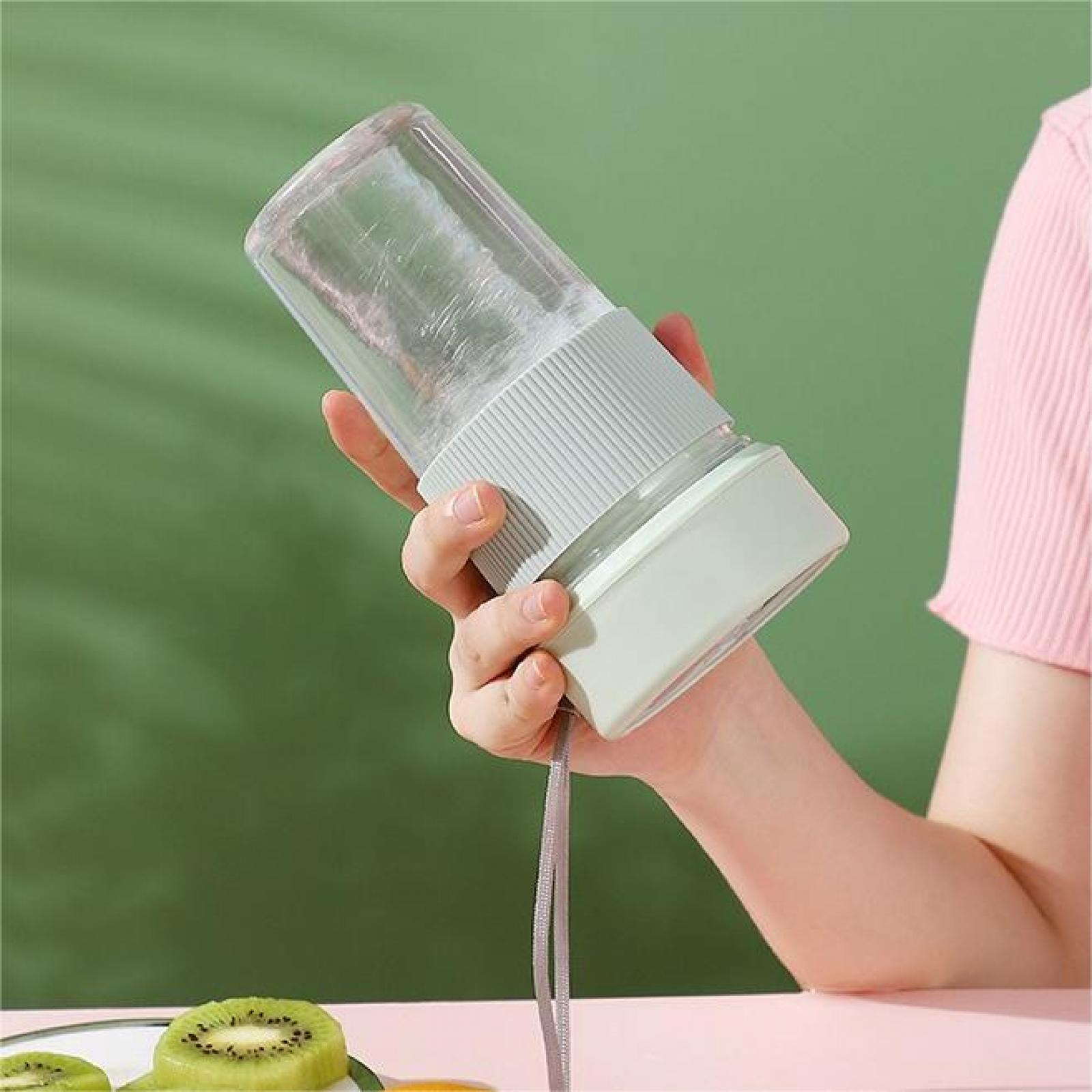 Die Hand einer Person, die einen Mixer mit grünem Sockel und einem transparenten Behälter hält, grüner Hintergrund, rosa Hemd.
