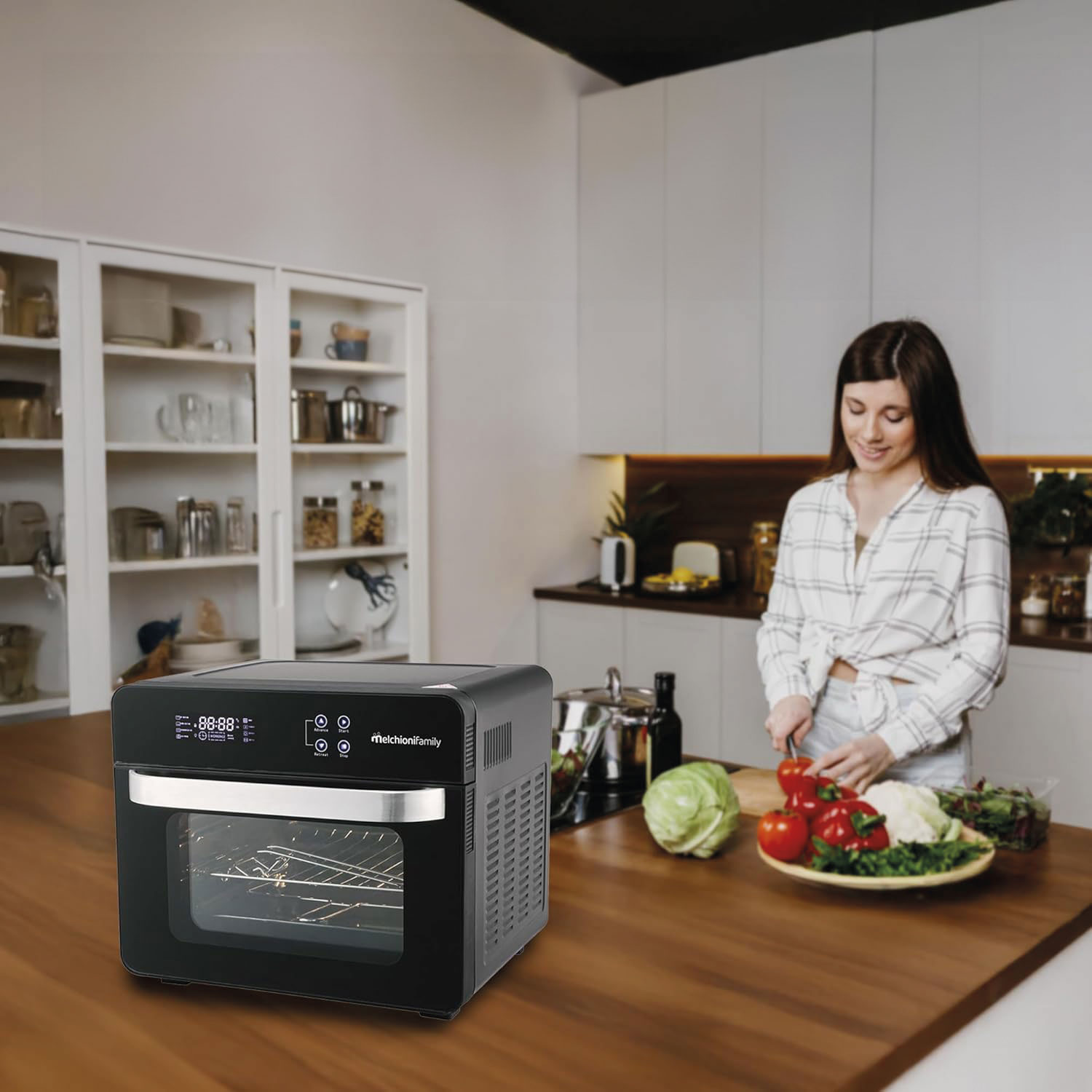 Una mujer corta verduras en una cocina. Una freidora de aire está sobre una encimera de madera. Armarios y estantes de cocina en el fondo.