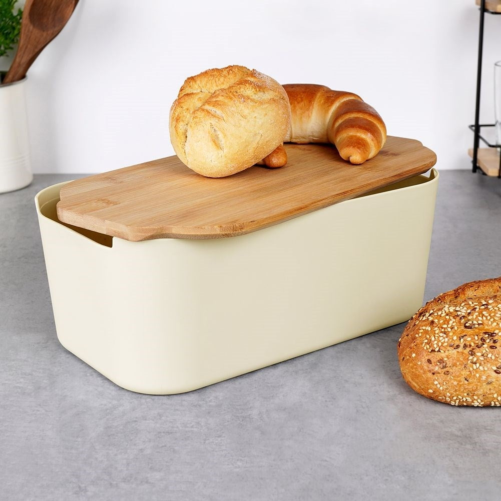 Un portapane color crema con coperchio in legno contiene pane e croissant. Una pagnotta è a destra su un piano grigio.