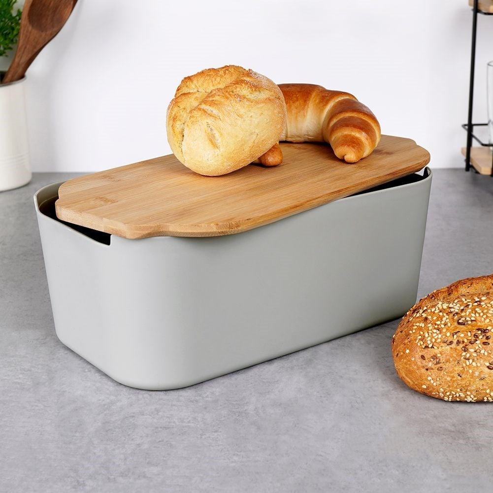 Un portapane grigio con coperchio in legno contiene un panino rotondo e un croissant. Una pagnotta intera è accanto.