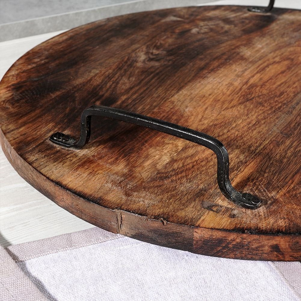 Primer plano de una bandeja redonda de madera con asa metálica. La bandeja es marrón y tiene vetas visibles. El asa es negra.