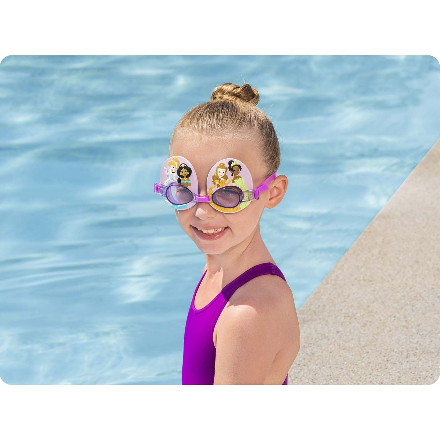 Una niña con gafas de natación sonríe. Las gafas muestran princesas de Disney. Lleva un bañador morado, de pie junto a una piscina.