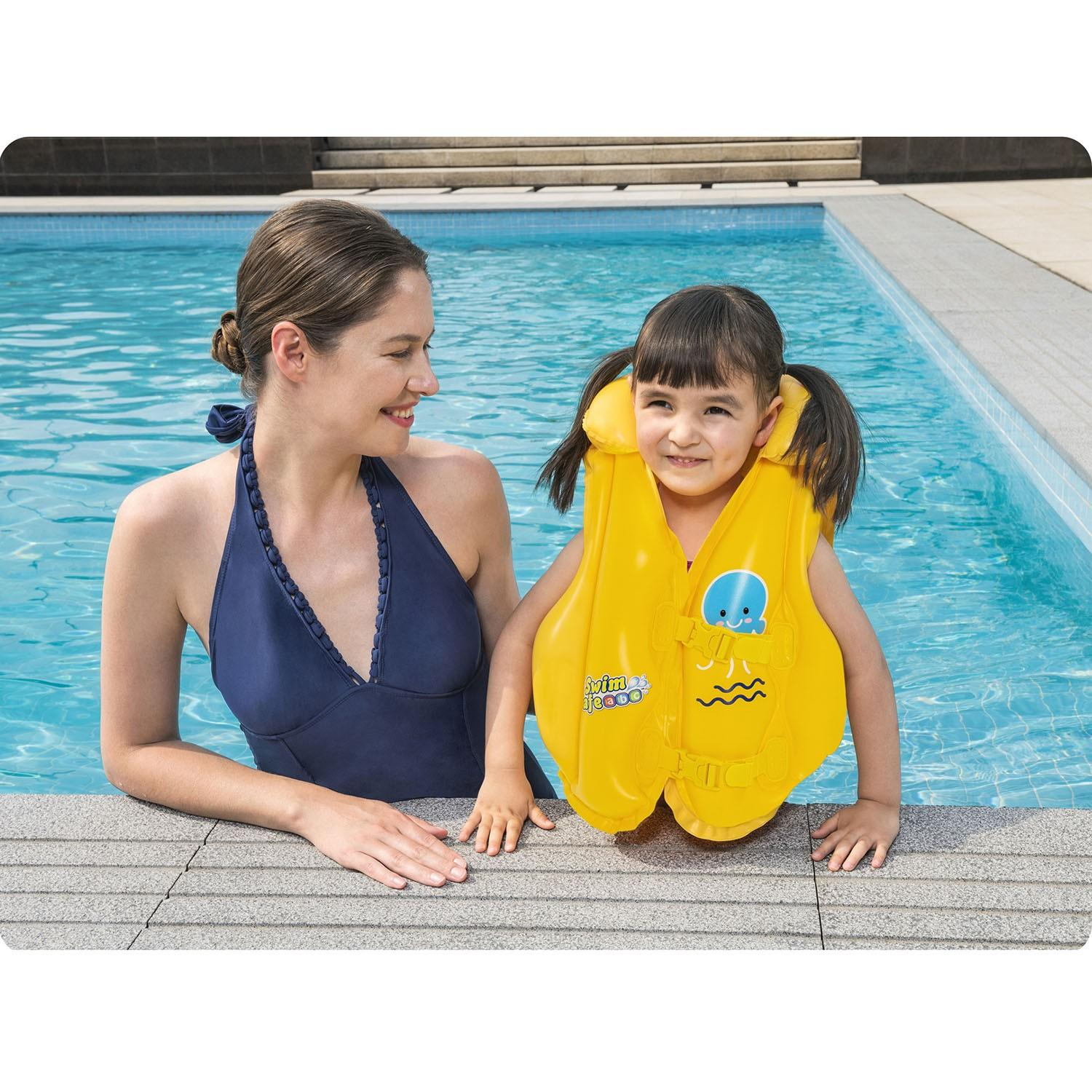 Una mujer y una niña en una piscina. La niña lleva un chaleco salvavidas amarillo. Ambas sonríen. Agua azul y piscina las rodean.