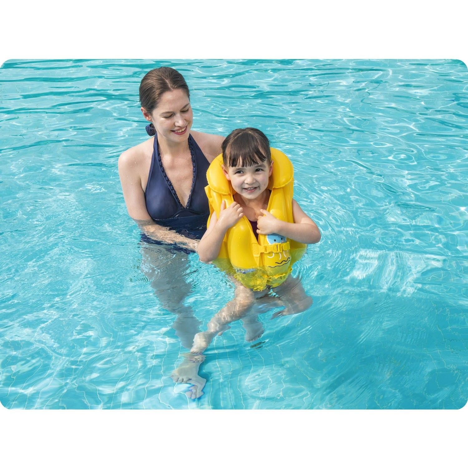 Una mujer y una niña en una piscina. La niña lleva un chaleco salvavidas amarillo. La mujer sostiene a la niña.