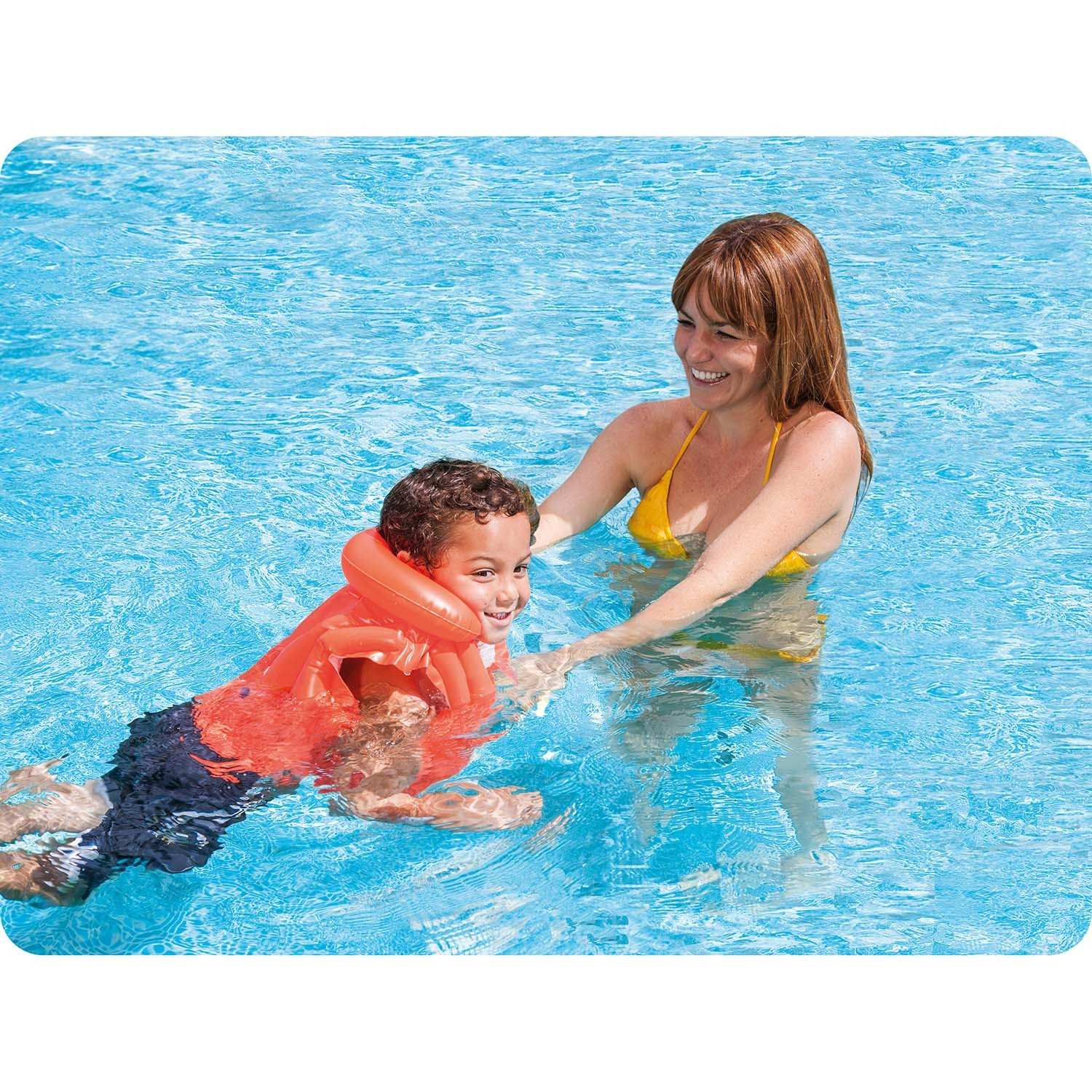 Una mujer en bikini amarillo ayuda a un niño con chaleco salvavidas a nadar en una piscina. Ambos sonriendo.