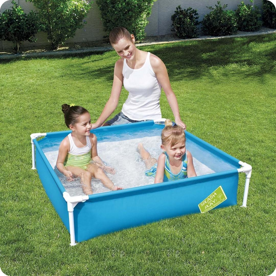 Dos niñas juegan en una pequeña piscina azul mientras una mujer observa en un césped verde. La piscina tiene un letrero de "Splash Play".