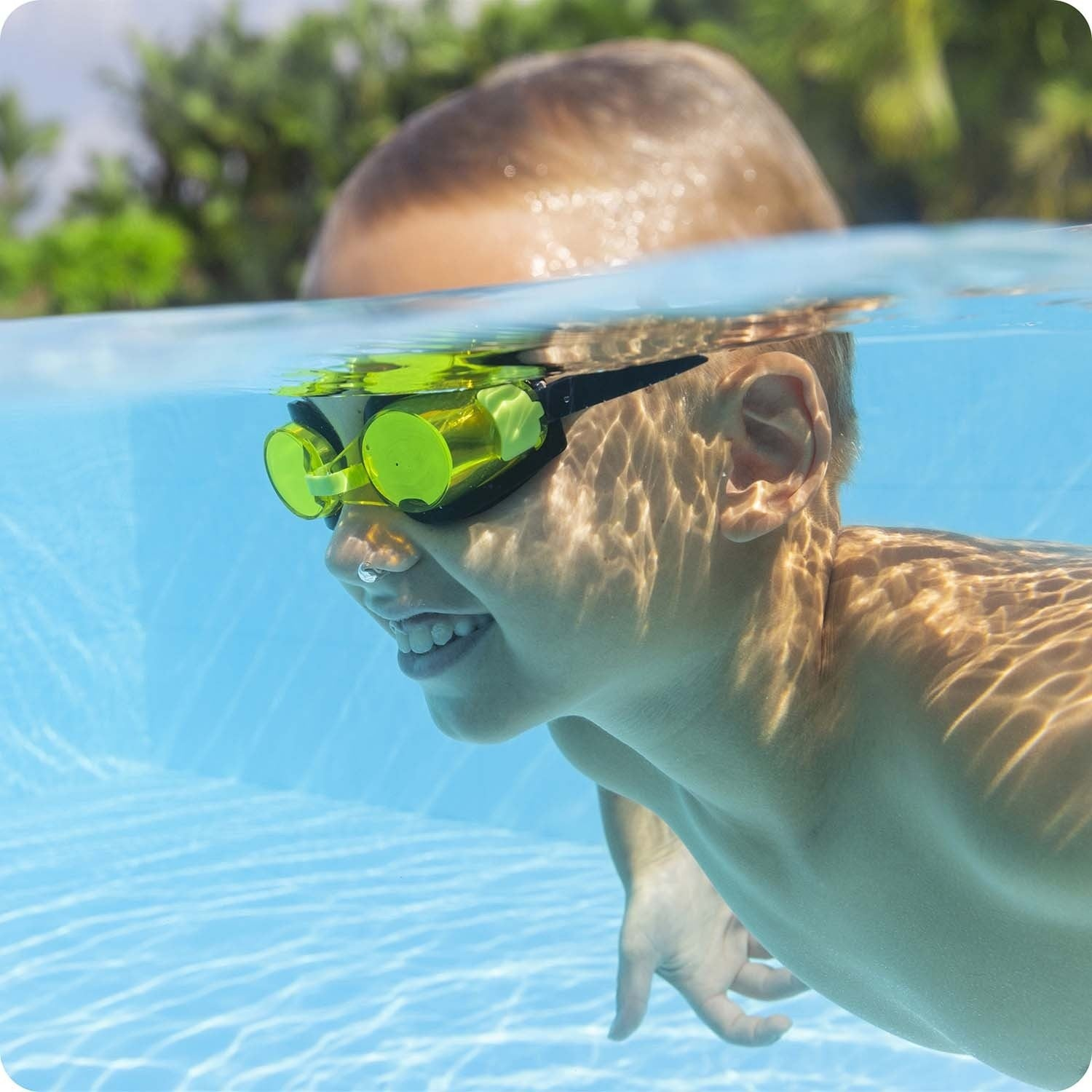 Un niño con gafas amarillas nada bajo el agua. La luz del sol brilla sobre su piel y el agua. Se ven árboles verdes al fondo.