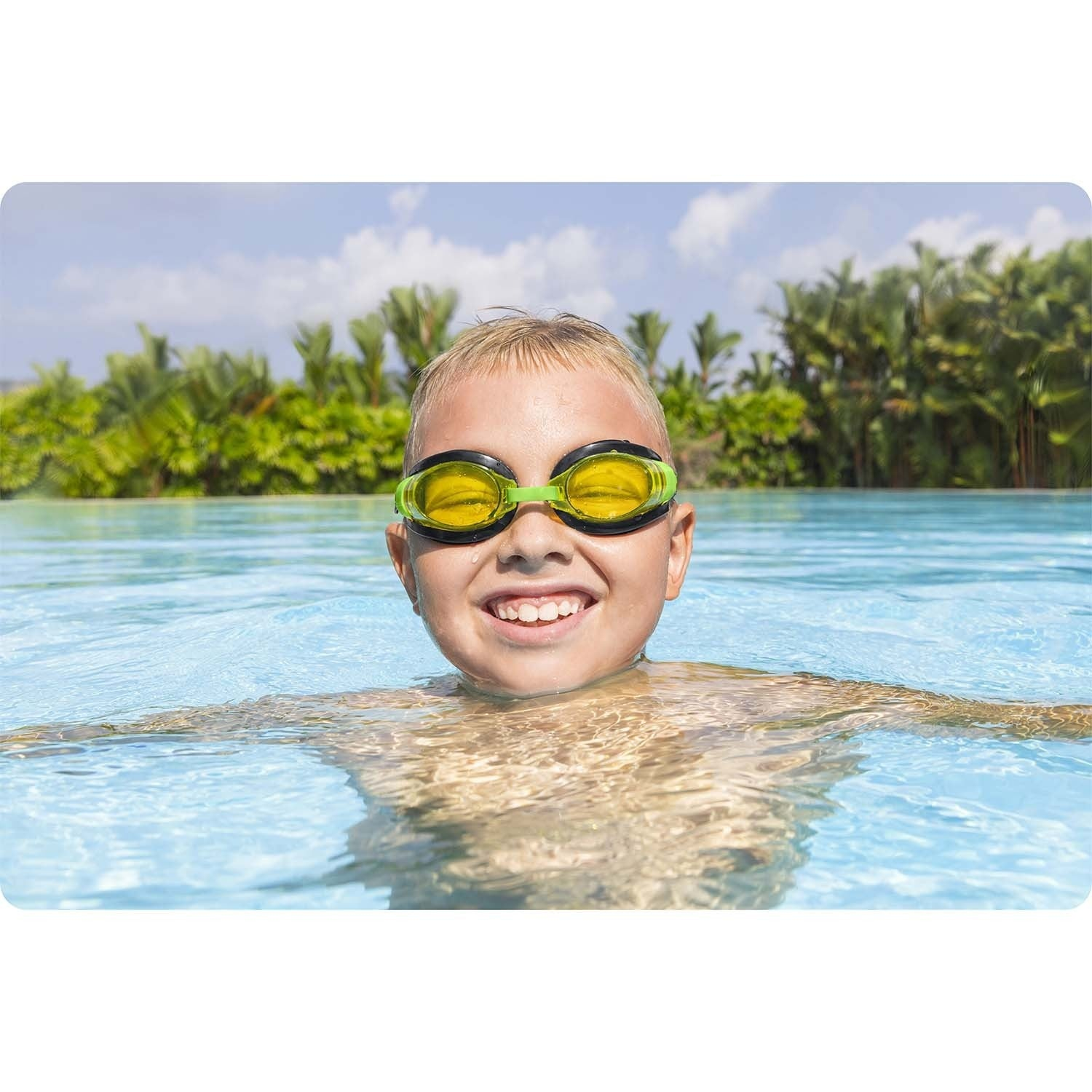 Un niño rubio con gafas de natación sonríe en una piscina. Palmeras y cielo azul en el fondo.