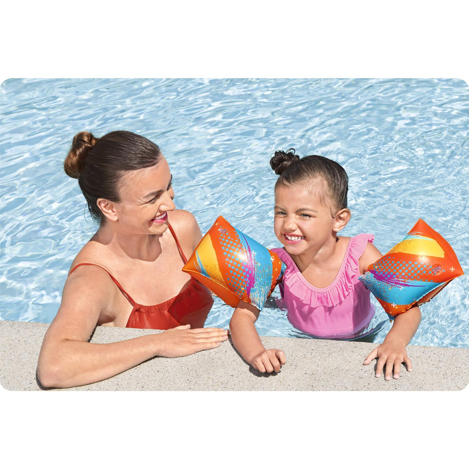 Una mujer y una niña en una piscina. La niña usa flotadores y un traje de baño rosa, sonriendo. La mujer sonríe también.