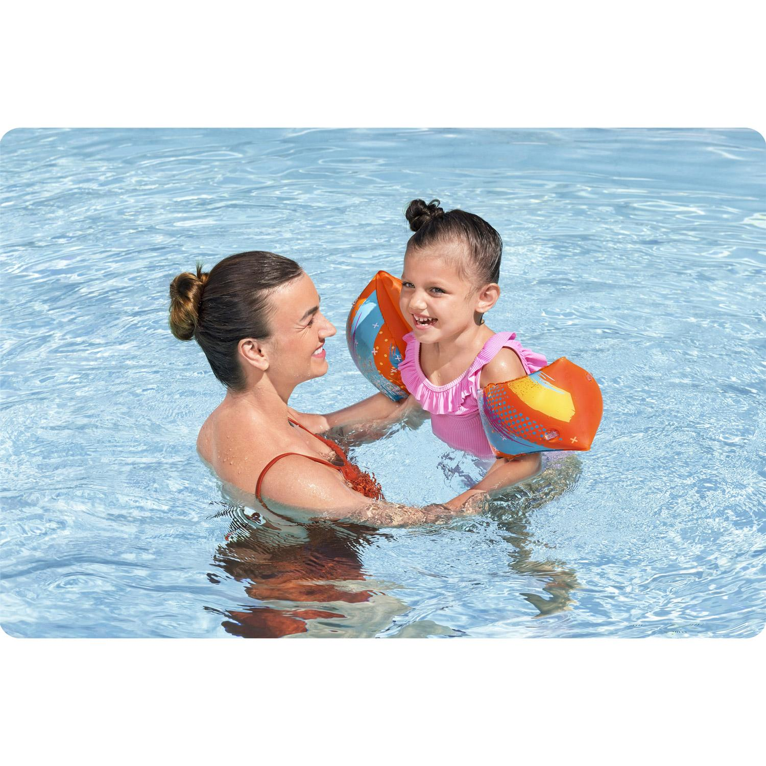 Una mujer y una niña están en una piscina. La mujer sostiene a la niña, que lleva flotadores y un traje de baño rosa.