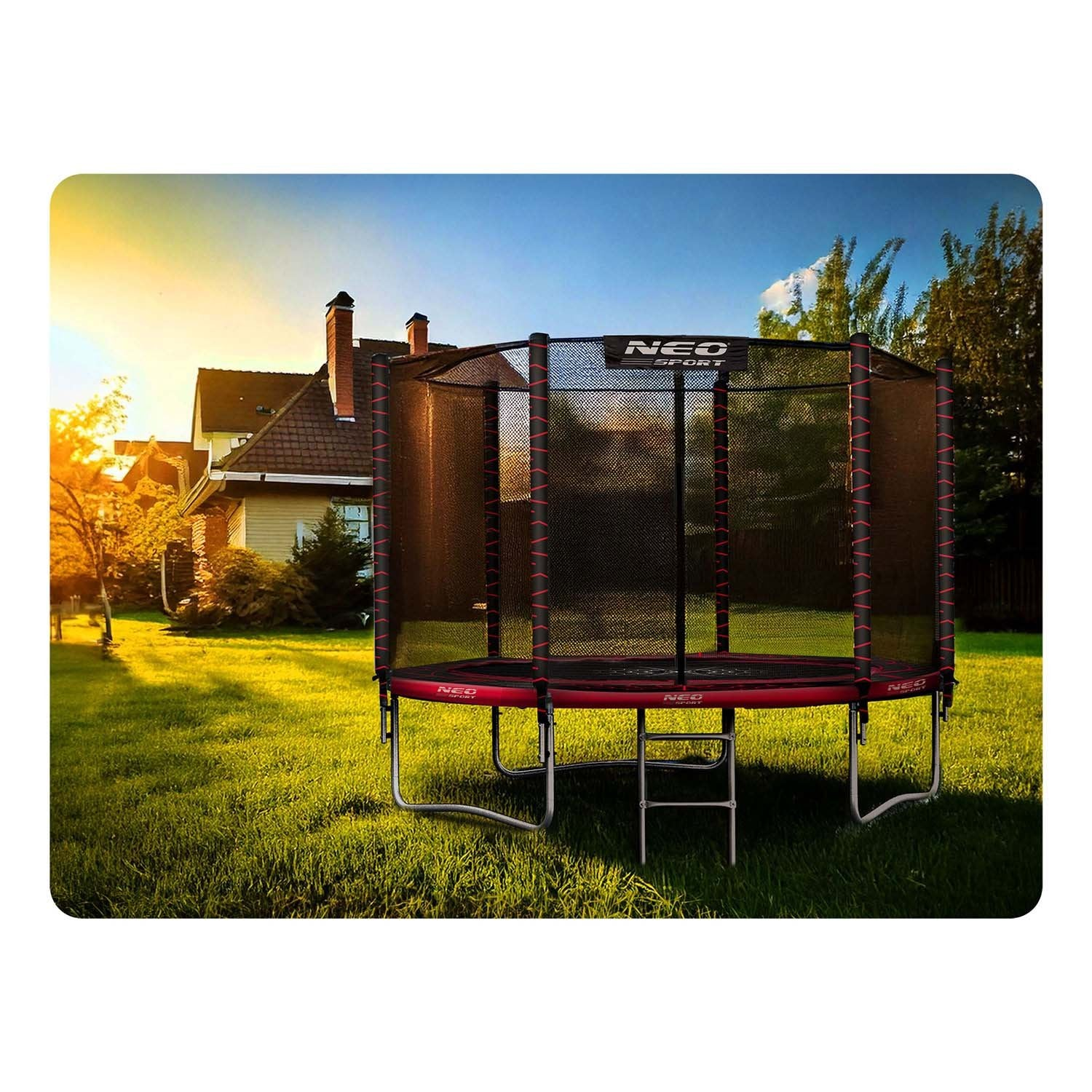 Un trampolín se encuentra en un jardín cubierto de hierba. Al fondo hay una casa con árboles y un cielo azul. El trampolín es rojo y negro.