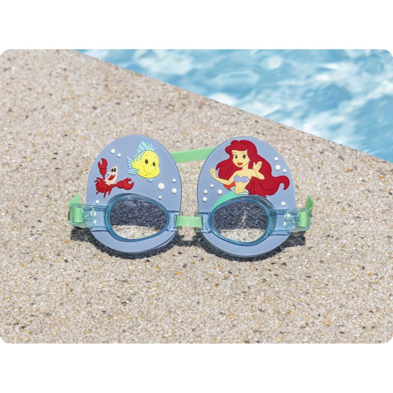 Gafas de natación con montura azul, con Ariel, un cangrejo y un pez amarillo. Descansan sobre una superficie de piedra, con una piscina de fondo.