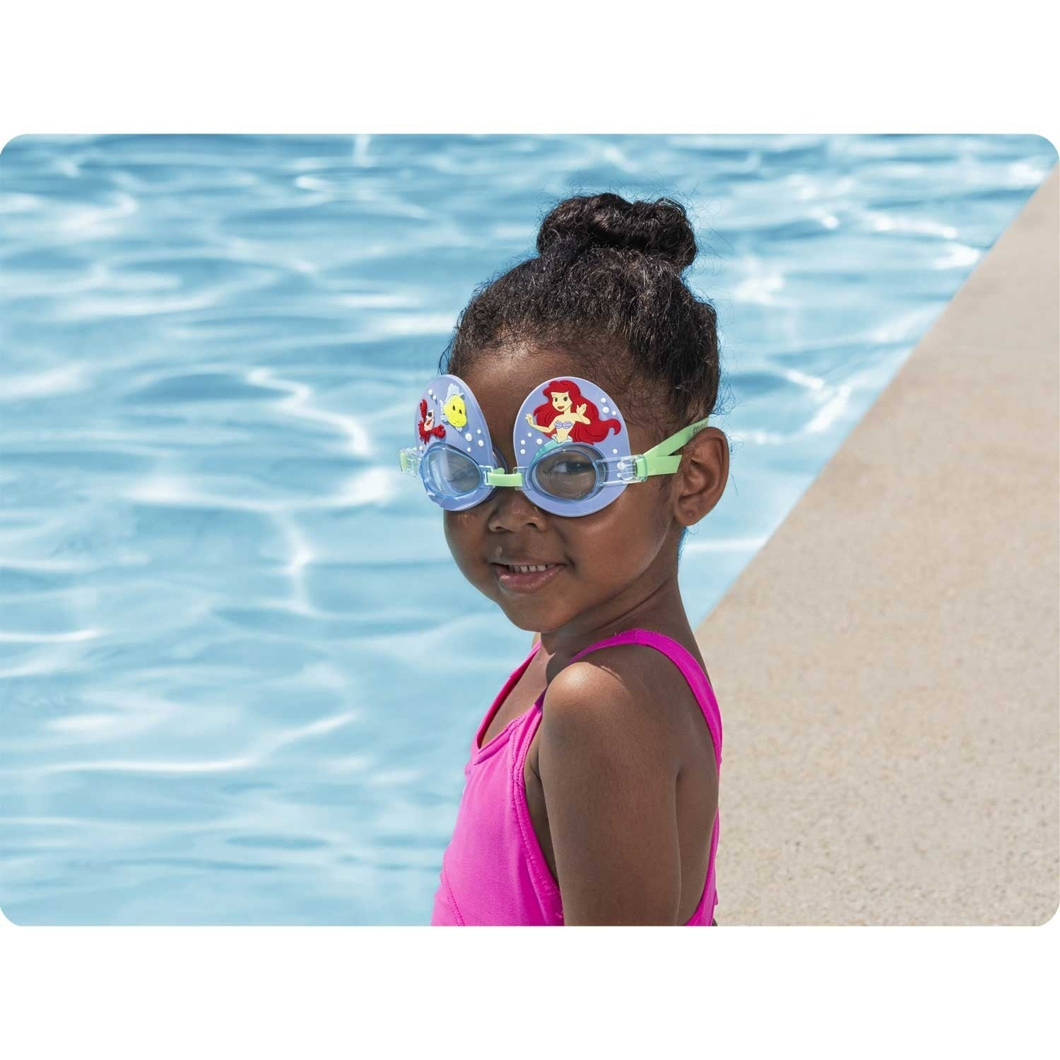 Una niña con gafas de natación con Ariel, en un traje de baño rosa. Está junto a una piscina, sonriendo.
