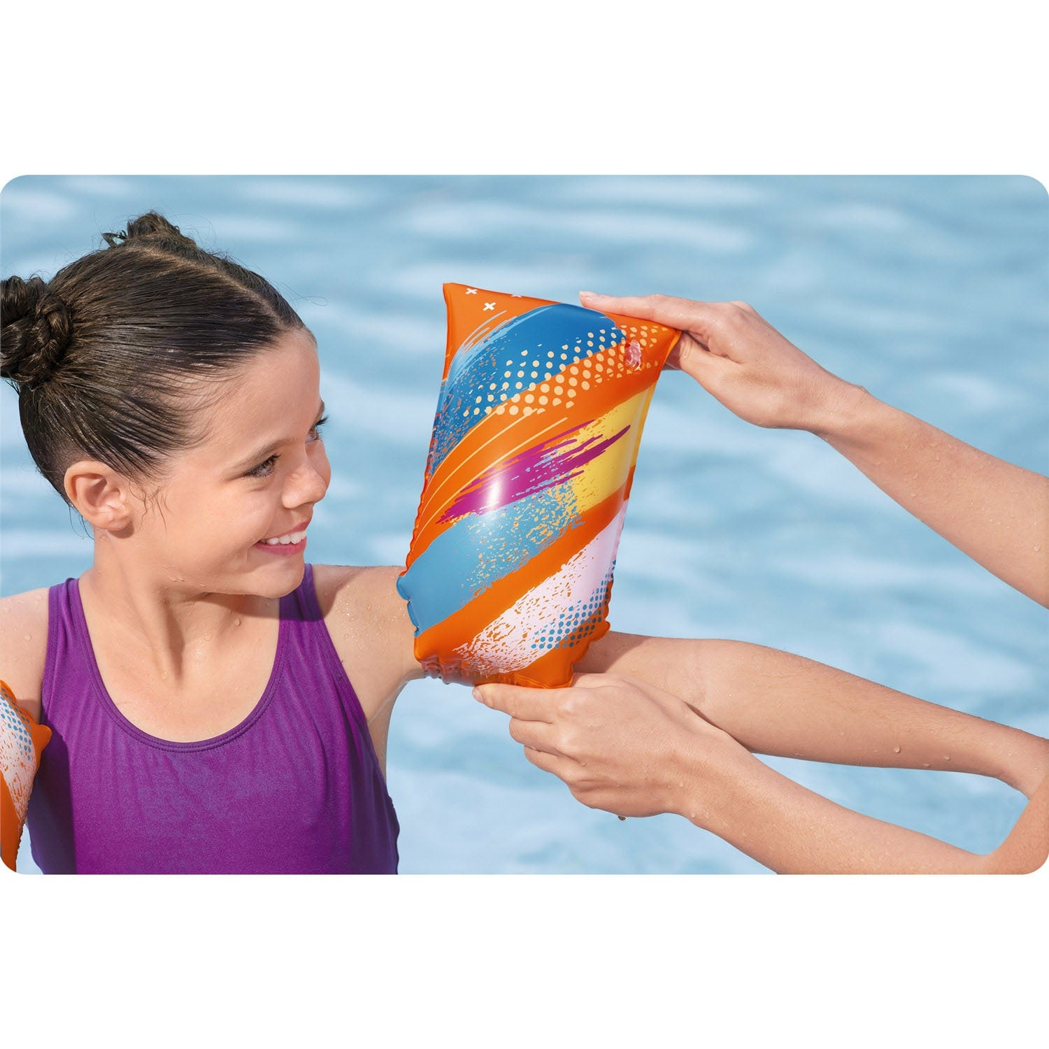 Una niña sonríe mientras alguien le ayuda a ponerse un flotador de brazo colorido. El fondo muestra una piscina.