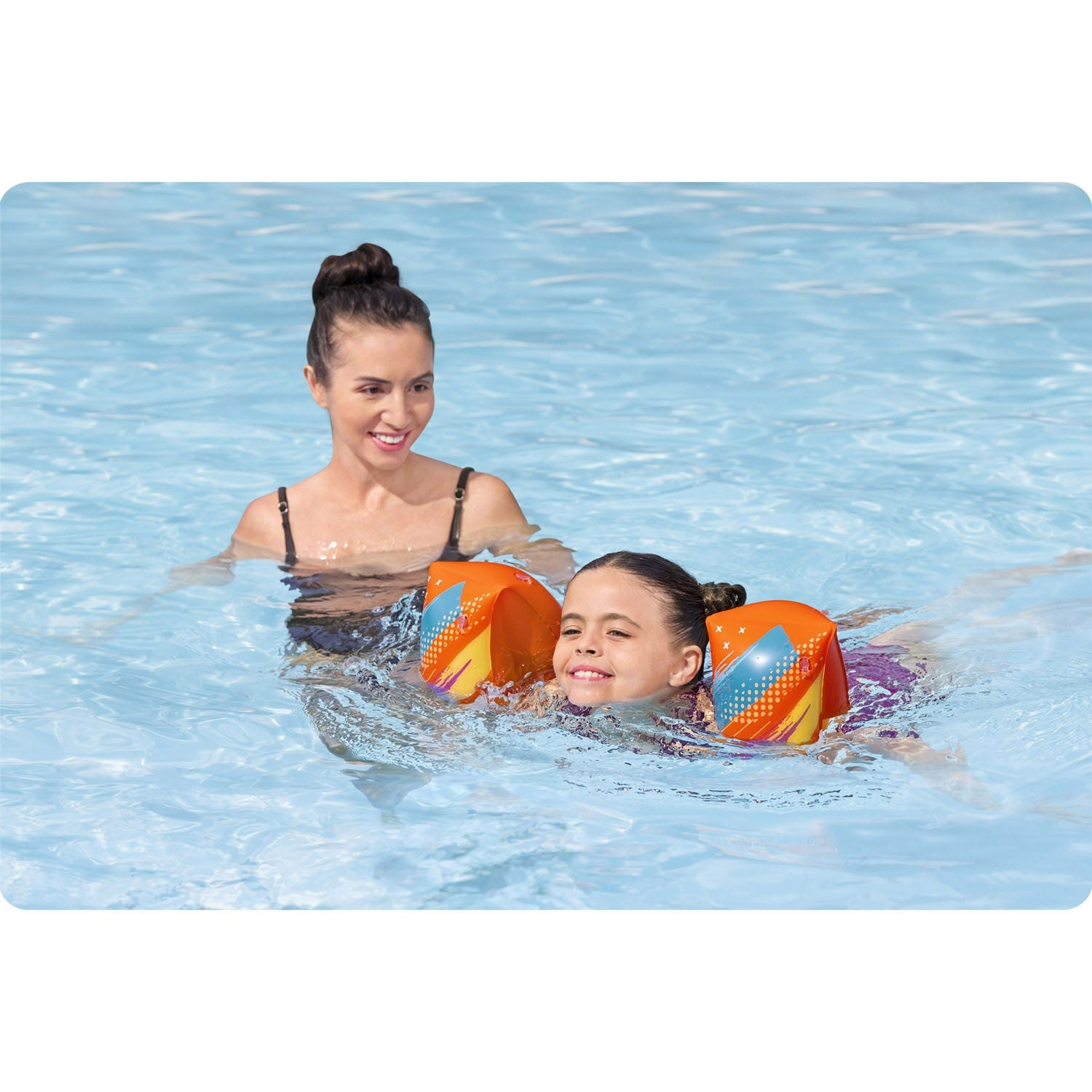 Una mujer y una niña están en una piscina. La niña lleva flotadores en los brazos. La mujer sonríe. El agua es azul.