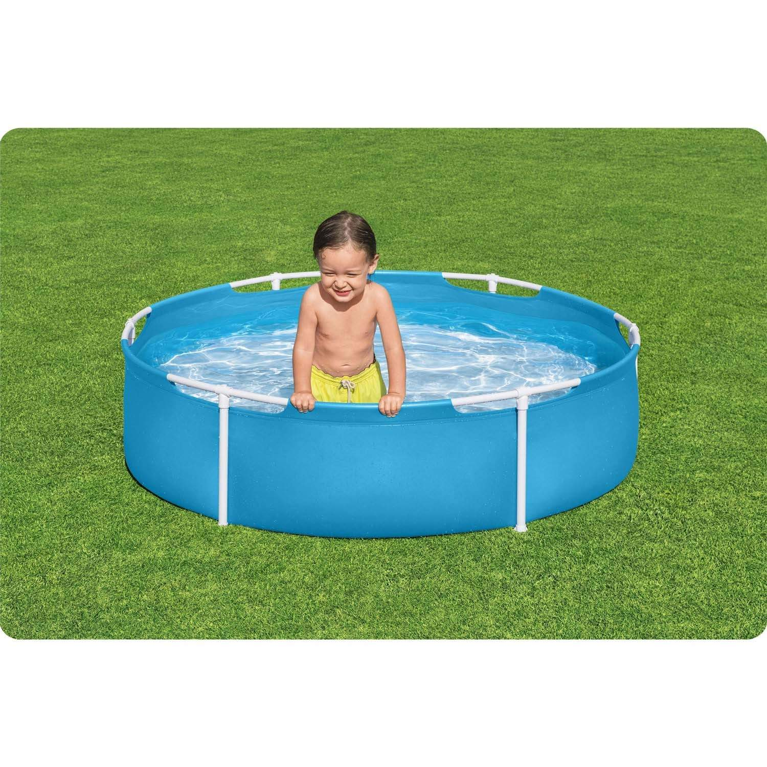 Un niño con pantalones cortos amarillos está en una piscina azul y redonda. La piscina está sobre hierba verde. El niño sonríe, mirando al espectador.