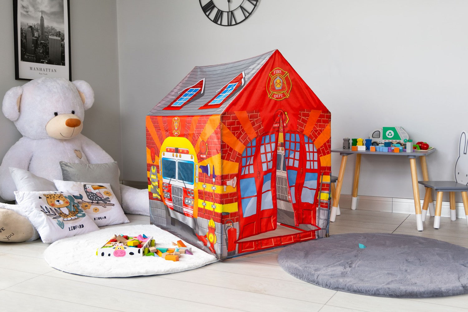 Una sala de juegos infantil con una casa de bomberos, un gran oso de peluche y una mesa pequeña con juguetes. Un reloj está en la pared.