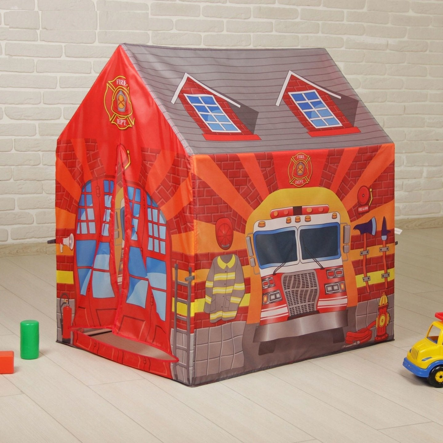 Una tienda de campaña de bomberos de juguete con fachada roja, ilustración de camión de bomberos y techo. Incluye bloques y un coche de juguete amarillo.