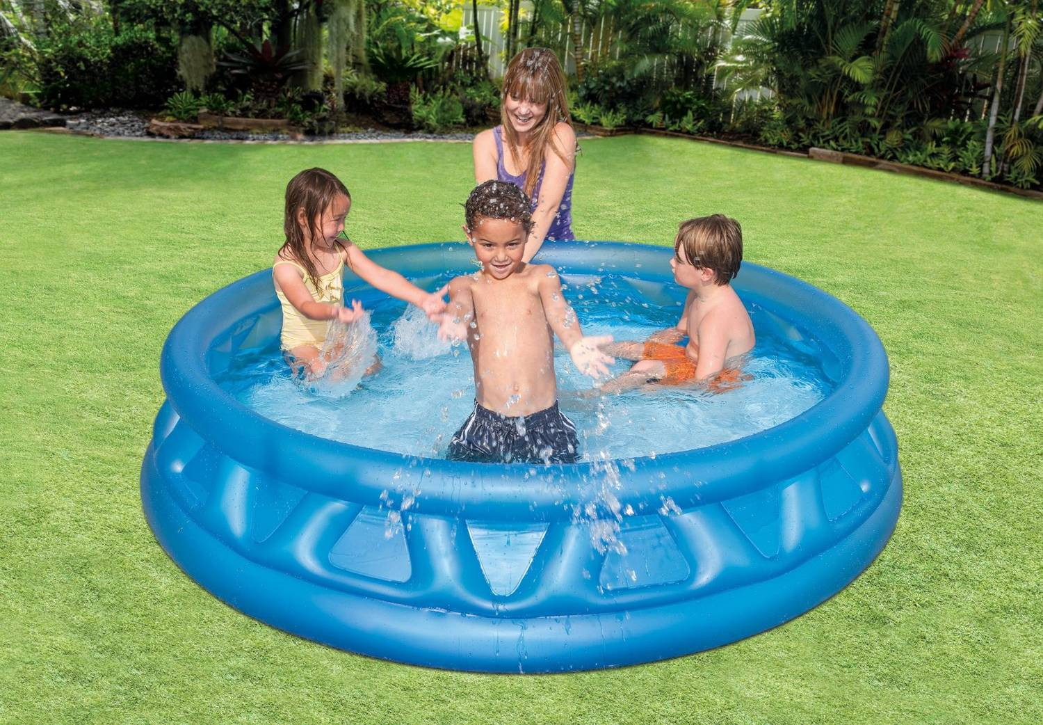 Cuatro niños y una mujer juegan en una piscina inflable azul sobre césped verde. Se ven salpicaduras de agua.