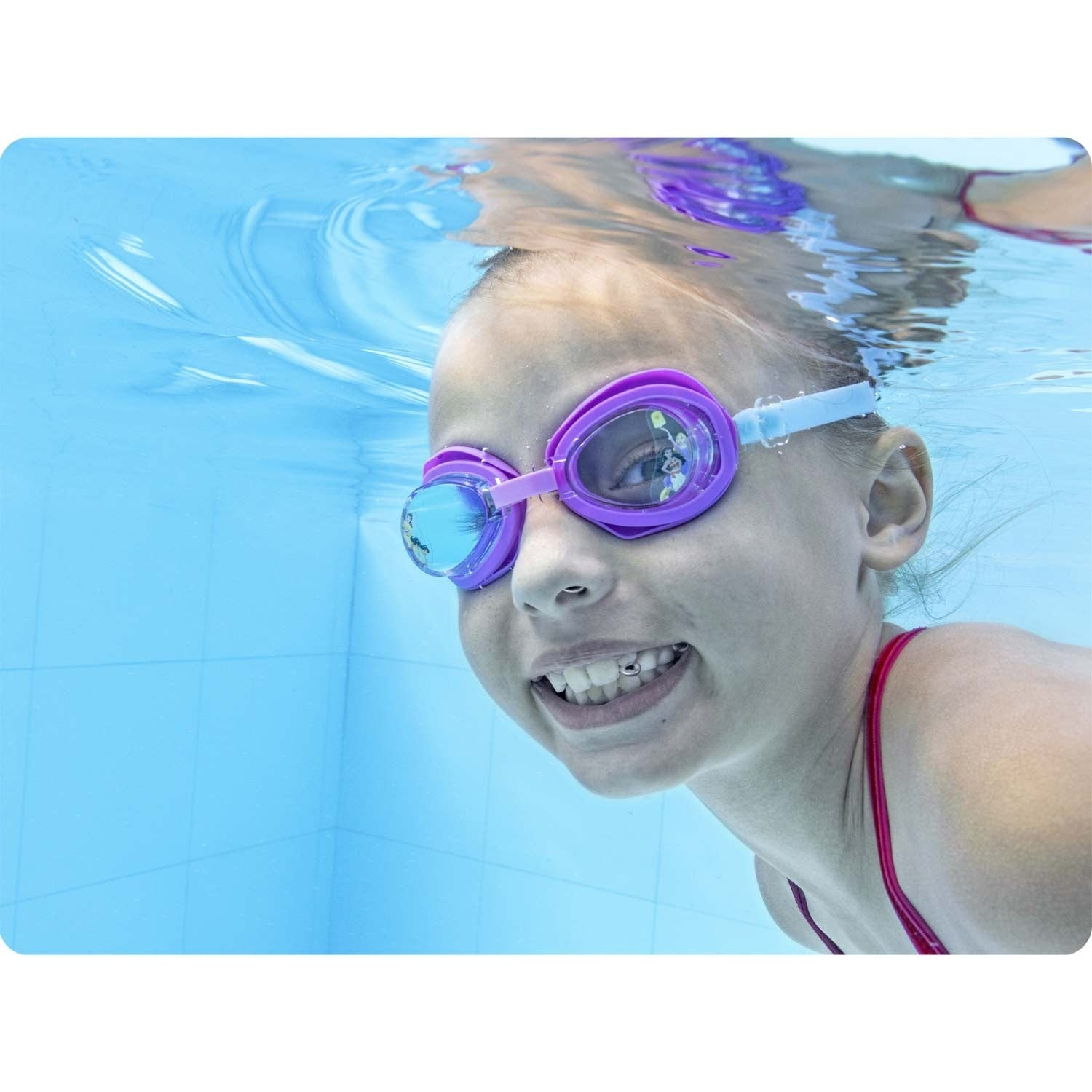 Una niña bajo el agua, con gafas moradas. Sonríe, con agua alrededor. El fondo es una pared de azulejos azules.