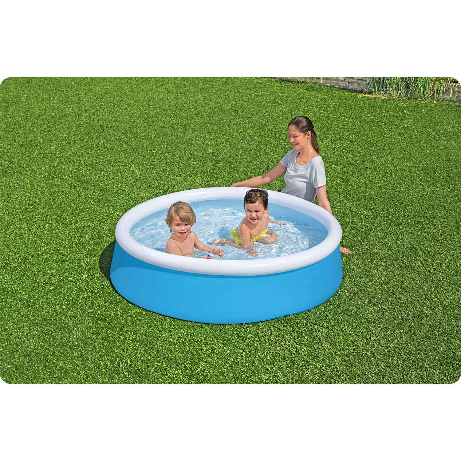 Dos niños pequeños en una piscina inflable azul con una mujer cerca sobre césped verde. La piscina tiene bordes blancos y azules.