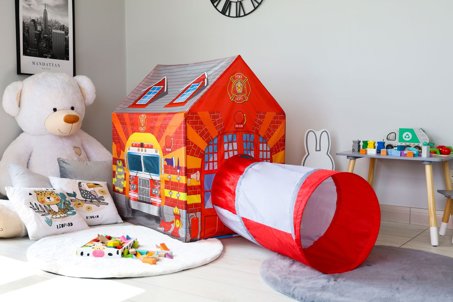 Una stanza giochi con una tenda dei pompieri, un orsacchiotto, giocattoli e un tunnel. La scena è luminosa e colorata, per i bambini.