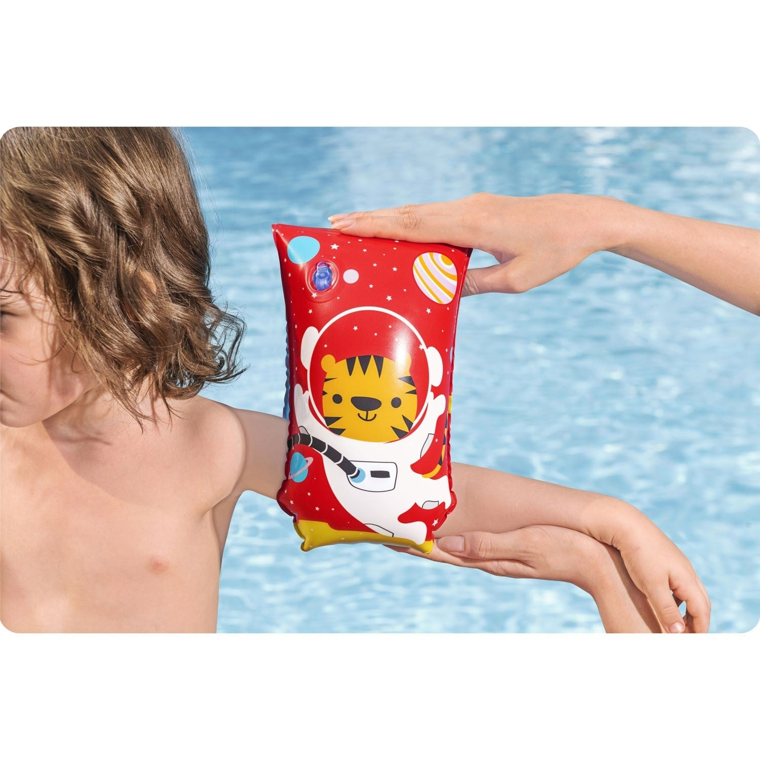 Un niño en una piscina con un flotador de brazo de astronauta rojo sostenido por una mano. El flotador tiene un diseño de tigre.
