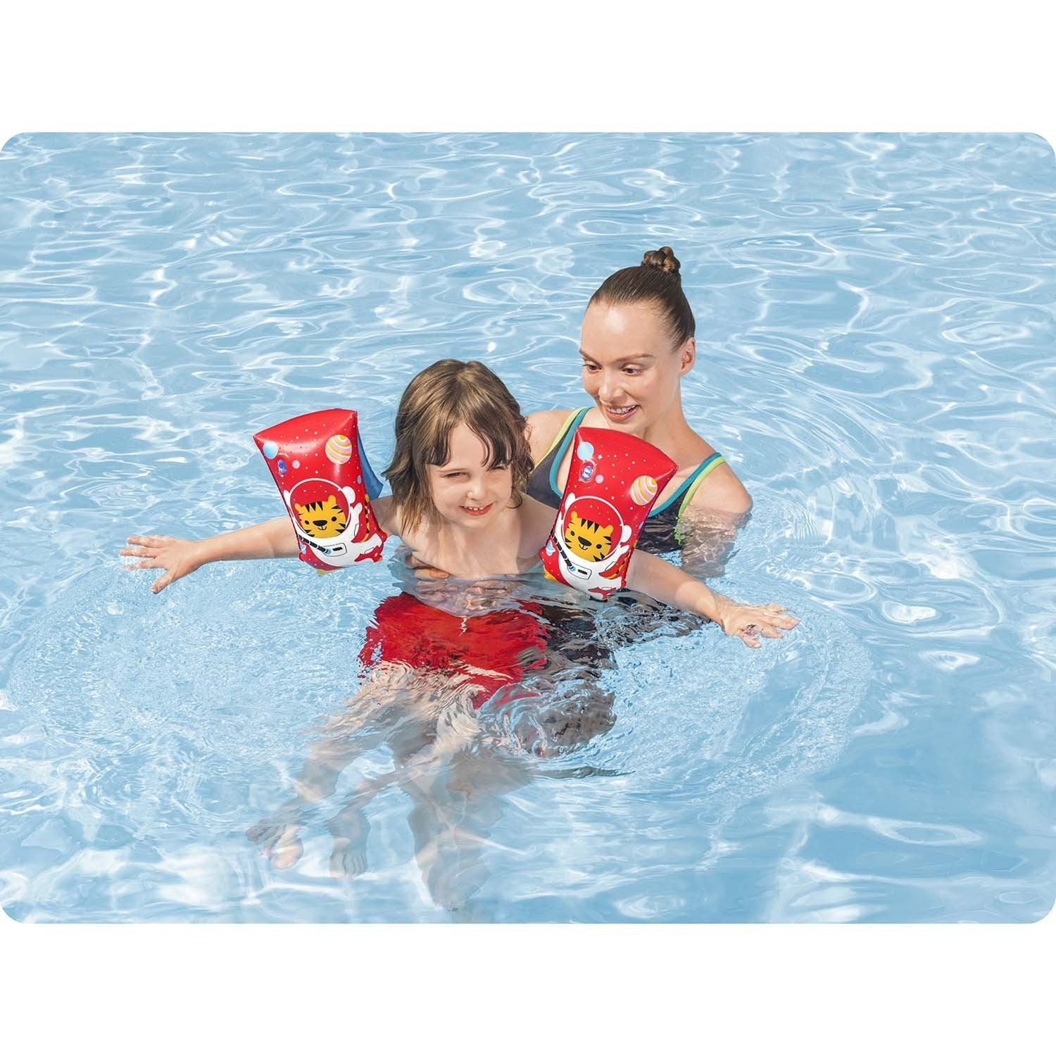 Una mujer sostiene a un niño en una piscina. Ambos usan flotadores de brazo. El niño en pantalones cortos rojos, la mujer en traje de baño. El agua es azul.