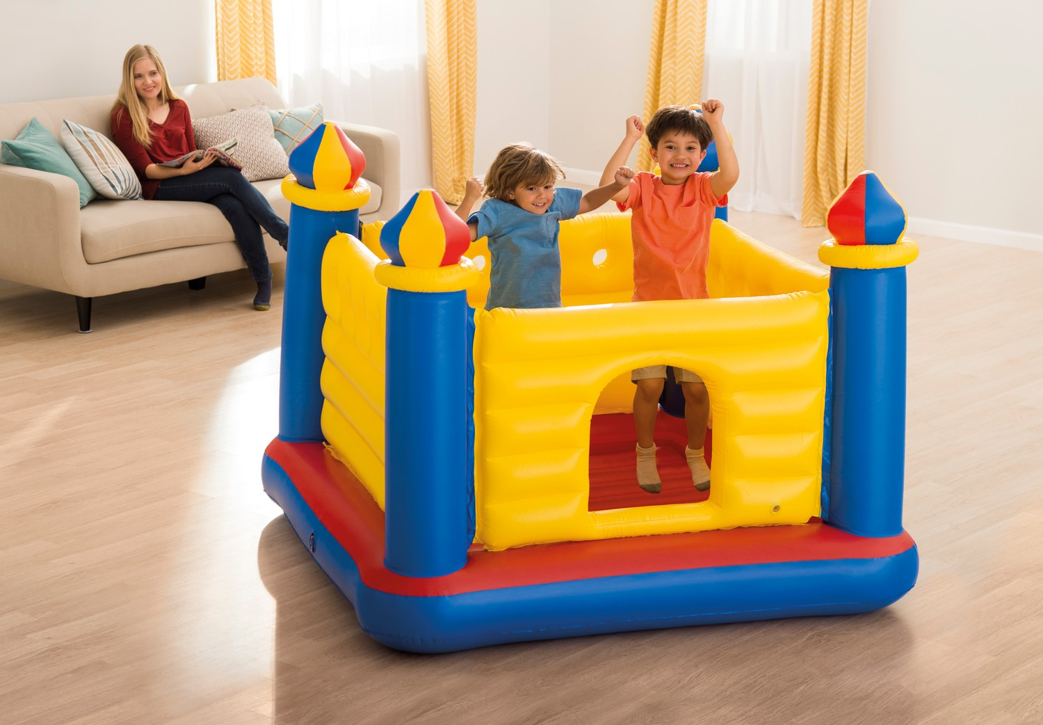 Dos niños saltan en un castillo inflable colorido. Una mujer lee en un sofá en el fondo.