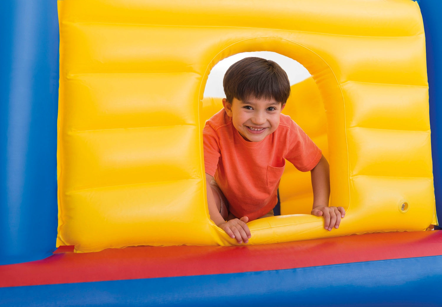 Un niño sonriente con una camisa naranja se asoma a través de una estructura inflable amarilla. Se ve azul, rojo y amarillo.
