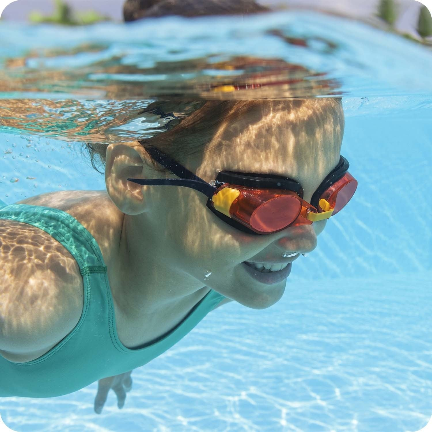 Una niña nada bajo el agua con gafas. Sonríe, el agua la rodea. La luz se refleja en su rostro y en el agua de la piscina.