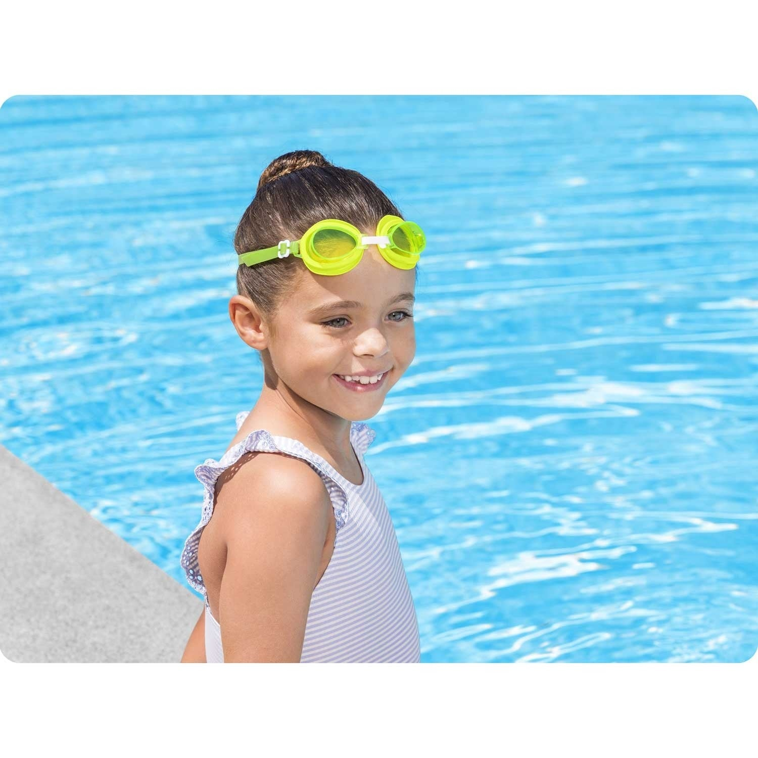 Una niña con gafas de natación sonríe junto a la piscina. Lleva un traje de baño a rayas. El agua de la piscina es azul.