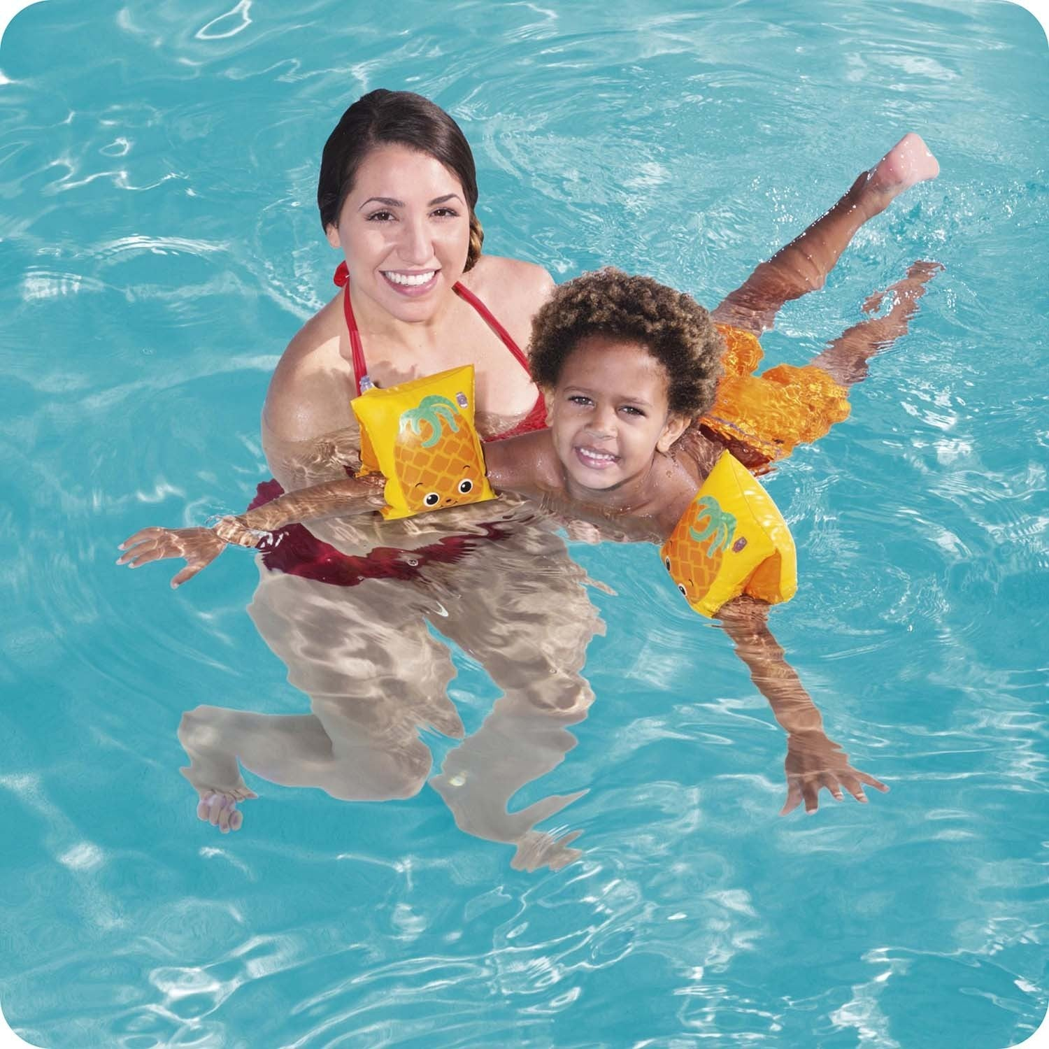 Una mujer sostiene a un niño en una piscina. Ambos usan flotadores de brazo. El niño usa pantalones cortos naranjas. Están sonriendo a la cámara.